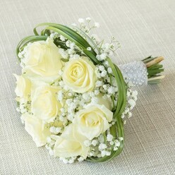 This elegant bouquet beautifully captures the spirit of Tufnell Park's quiet residential charm, delicately arranged and photographed atop a woven beige fabric that highlights its nuanced palette. Creamy white and pale yellow roses-eight to ten in full, velvety bloom-form the core of the bouquet, their petals opening from tight buds to sumptuous, open blossoms with a gentle radiance reminiscent of morning light filtering through Victorian terraces on Campdale Road. Interspersed throughout, clusters of cloud-like baby's breath bring a weightless, romantic quality, their miniature white florets lending both visual softness and a hint of delicate scent you might associate with early spring strolls through Tufnell Park's pocket parks. Spirals of vivid green foliage, perhaps bear grass, thread dynamically among the blooms, curving over and around them in graceful sweeps that create a balanced yet contemporary silhouette, echoing the blend of tradition and innovation seen in local floristry. The stems are elegantly cinched together with a satiny grey ribbon, its surface sparkling with multiple rows of pearl beads-each one adding a note of tactile luxury and anticipation for a special day. The fresh-cut stem ends peek out proudly, a reminder of the bouquet's freshness and handcrafted origin. Ideal as a statement piece for a wedding at a local venue or a celebration among friends, this arrangement radiates understated opulence, crafted with the discerning eye and gentle touch found at Tufnell Park's favourite flower shop.