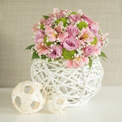 This sophisticated wedding table centerpiece draws inspiration from the calm, community-driven atmosphere of Tufnell Park, presented in a luxuriously abundant floral display atop a pristine white surface. Opening with a foundation of lush, fully-opened pastel pink and lavender roses, the bouquet instantly suggests a mood of tender celebration-each bloom exuding the cool softness of velvet petals familiar to anyone who's wandered through Tufnell Park's seasonal florists. Pink alstroemeria blooms, with their trumpet-shaped petals streaked in gold and cerise, intertwine thoughtfully with the roses, enhancing the arrangement's textural play. Bright lime green 'Kermit' button chrysanthemums add a buoyant pop of contemporary energy, offset by fine clouds of airy baby's breath that lend lightness and gentle movement. Rich, forest-green leaves and foliage bridge the tones, providing both a visual anchor and a sense of garden freshness akin to walks through the gardens near Dartmouth Park Hill. The flowers rise from a unique white vase, its intricate woven pattern resembling delicate basketwork and embodying both rustic and modern influences. Nearby, light-toned decorative spheres-fashioned from woven rattan-echo the vase's artistry and add continuity, inviting the eye to linger. Diffuse natural light washes over the soft beige backdrop, bathing the scene in an ambiance that feels calm and reassuring. This composition, crafted with imagination and local expertise, feels equally suited to special events or elegant dining in homes across Tufnell Park and Kentish Town, bringing a sense of quiet confidence and refined joy.