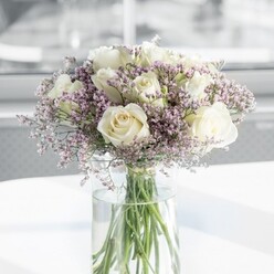 In this photograph, a beautifully balanced bouquet of creamy white roses and soft lilac statice is presented in a tall, clear glass vase, capturing a mood of calm sophistication that feels right at home in Tufnell Park. The arrangement is almost perfectly spherical, with about a dozen white roses forming a generous, rounded canopy. Each bloom is at that ideal stage between bud and full flower, the petals gently opening to reveal layered hearts that look plush and velvety. Between these classic roses, wisps of tiny lavender-tinted statice appear like delicate lacework, filling the spaces without crowding them, and introducing a gentle haze of colour that plays between cool lilac and muted purple. The stems, visible through the cylindrical vase, are neatly trimmed and arranged so they lean gracefully, their fresh green tones deepening where they meet the clear water. The vase rests on a smooth, white surface that bounces back the daylight, adding a soft glow to the underside of the bouquet and enhancing the sense of cleanliness and modern style. Behind the arrangement, the background is blurred into a wash of pale, neutral tones, suggesting an airy room, possibly a contemporary flat near Tufnell Park Fields or a bright office tucked away off Junction Road. The natural light seems to stream in from one side, kissing the tops of the petals and casting the faintest of shadows, which gives the bouquet depth and dimension. The feeling is one of understated grace-an arrangement well suited to a quiet morning celebration, a wedding breakfast table, or a respectful, comforting gift delivered across the local neighbourhood. The image conveys the careful eye and gentle touch of a florist who understands how to let each flower breathe, while still holding the whole composition in perfect harmony.