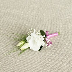 Elegantly placed on a pale beige, linen-like cloth, this handcrafted boutonnière feels like a calm moment before a wedding begins in Tufnell Park, perhaps on a tree-lined street near Tufnell Park Road. The central focus is a single, fully opened white lisianthus flower, its petals gently ruffled and layered, forming a soft, rounded cup that looks almost weightless against the neutral background. Just beside it, two graceful lisianthus buds, still closed in shades of cream and pale green, lean slightly towards the bloom, adding a sense of movement and anticipation, as if capturing that tender pause before vows are spoken. Scattered thoughtfully around the main flower are tiny clusters of white baby's breath, each minuscule blossom adding a whisper of texture and creating an airy halo around the focal bloom. Fine, grass-like strands of greenery stretch out to the left, slender and dark green, giving a natural, slightly wild edge to an otherwise refined design, while a broader, deep green leaf sits quietly at the back, providing structure and a gentle shadow that defines the white petals. At the base, the stems are bound with a vivid fuchsia pink satin ribbon, its surface smooth and slightly glossy, tied in a neat little bow that injects a playful yet chic accent of colour. The lighting is soft and flattering, revealing each detail without glare, making it easy to imagine this boutonnière pinned to a groom's jacket at a Town Hall ceremony or prom night near Hampstead Heath. The composition conveys elegance, freshness, and a sense of intimate celebration, perfectly suited to local couples seeking understated refinement with a pop of personality.