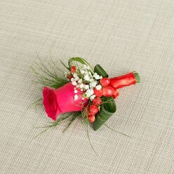 Set against a neutral, woven beige background that resembles softly lit linen, a handcrafted buttonhole from a Tufnell Park florist captures the essence of a romantic North London celebration. The composition centres on a single, deep fuchsia pink rose, its petals dense and velvety, curling inward at the core and gradually opening at the edges, as if in mid-breath before the ceremony begins. Surrounding the rose, clusters of white baby's breath create a gentle, mist-like frame, their tiny blossoms offering a sense of purity and lightness that softens the strong colour of the bloom. Between these airy flowers, small scarlet hypericum berries glisten, their smooth, rounded forms adding both texture and a subtle hint of festivity, ideal for weddings held near the leafy streets between Tufnell Park and Kentish Town. Delicate, fern-like greenery extends outward in fine lines, contrasting with the broader, darker green leaves that have been expertly curved to support the main rose, giving the arrangement a sculpted yet natural look. The stems are gathered tightly and wrapped in a ribbon of rich red satin, the bow small, precise, and slightly reflective, suggesting a careful, practiced touch. Soft, diffused daylight falls across the fabric and the boutonniere, creating gentle shadows that highlight its contours and layers. The overall mood is elegant and heartfelt, perfectly suited to a groom or groomsman arriving at a local registry office or family gathering, carrying with them this small but meaningful token of colour, fragrance, and celebration.