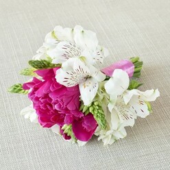 A small, meticulously styled floral arrangement rests horizontally on a pale, linen-like fabric, its soft beige weave providing a calm, neutral backdrop for the vivid colours of the flowers. At the centre, a fully opened fuchsia peony steals attention, its countless ruffled petals forming a lush, rounded bloom with a rich, almost silky texture. Surrounding this striking flower are several white alstroemeria, or Peruvian lilies, their trumpet-shaped petals gently flaring outward. Each white petal is intricately patterned with fine burgundy streaks and freckles, adding a sense of delicate, handwritten detail to the arrangement. Slender spikes and clusters of fresh green buds weave in and out between the flowers, bringing a lively, linear contrast and a sense of new growth, much like the fresh greenery seen along Tufnell Park Road in late spring. The stems at the base are neatly gathered and tightly secured with a bright pink satin ribbon whose sheen echoes the vivid magenta tones of the peony, creating a cohesive and polished finish. The lighting is soft and even, highlighting the natural textures of petal, leaf, and fabric without harsh shadows, so every curve and fold feels calm and intentional. This compact design feels ideal as a boutonniere, corsage, or intimate hand-held posy for weddings and celebrations around Tufnell Park, from registry office ceremonies to relaxed gatherings at local cafes. The overall impression is one of modern romance, combining bold colour, fine detail, and a sense of effortless elegance shaped by an experienced local florist.