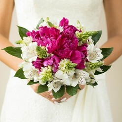 The image shows a tightly framed, intimate view of a bridal-style bouquet being held close to the waist of someone dressed in a white wedding gown, the subtle texture of lace or fine fabric forming a calm, neutral backdrop. Dominating the arrangement are voluptuous magenta and fuchsia peonies, each bloom packed with layers of ruffled petals that catch the light in soft highlights and shadows, creating a sense of depth and opulence. Interspersed through these showy flowers are white alstroemeria, also known as Peruvian lilies, with their slender petals and distinctive maroon flecks concentrated in the throat of each blossom, adding an almost painterly detail. Between the larger blooms, sharp, upright green buds and textured filler stems punctuate the design, contributing a lively, fresh-from-the-market feel that recalls early morning flower runs along nearby Camden and Holloway Road. Dark, glossy leaves curve around the outside, rounding off the bouquet into a balanced, almost perfectly spherical shape that sits comfortably in the hands. The person holding it has neatly shaped nails painted a gentle blush pink, reinforcing the soft, romantic mood. The focus is crisp on the flowers, while the background gently blurs, concentrating the viewer's attention on the vivid interplay of fuchsia, white, and green. This bouquet feels like a bespoke creation for a Tufnell Park wedding, ideal for a ceremony followed by a reception in one of the area's cosy venues, expressing modern romance with a hint of classic London elegance in every petal and leaf.