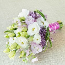 Radiating refined charm from a local Tufnell Park florist, this exquisite bouquet stretches diagonally across a pale linen canvas, the neutral backdrop allowing every colour and texture to glow in the soft, natural light filtering through a London window. The arrangement effortlessly marries tradition and contemporary elegance, with fully open snow-white Lisianthus flowers at its heart, their ruffled petals caressing buttery yellow-green centers in a gesture of gentle warmth. Around them, numerous slender Lisianthus buds-still in tight formation-whisper of new beginnings, while dusky lavender-pink stocks create tender pockets of scent and movement, reminiscent of a stroll through Tufnell Park's community gardens in full bloom. The bouquet's drama comes alive through deep purple allium, their spiky, star-like florets adding a sculptural, modern dimension just as urban parks provide striking green escapes. Weaving throughout are lively lime-green buds and sweeping strands of bear grass, interlacing with textured foliage to imbue a sense of depth and spirited vitality. All is gathered at the stems with a joyous burst of fuchsia pink satin ribbon, overlaid with pure white pearls-a nod to timeless sophistication and joyful festivity, perfect for a wedding morning or anniversary. Each element is thoughtfully chosen and composed, echoing the creative soul of Tufnell Park and bringing together the optimism, artistry, and celebration of connection that define this neighbourhood's most cherished events.