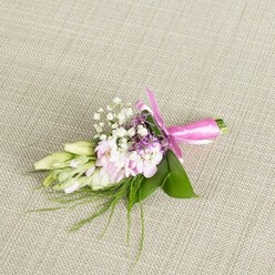 On a pale, finely woven linen surface that looks like the sort of relaxed yet refined fabric you might find in a Tufnell Park townhouse, a single floral boutonniere or petite corsage is carefully laid out, its details clearly visible. The flowers themselves form a compact oval, roughly the length of a gentleman's boutonniere, styled for a wedding, prom, or intimate celebration at a local venue near Tufnell Park Underground or the green edges of Parliament Hill Fields. At the centre, a cluster of soft light pink blooms-delicate, multi-petalled flowers reminiscent of miniature garden stocks or small ruffled roses-gathers in a loose, natural mound. Their petals have a velvety texture and a gentle gradation from near-white at the edges to a more noticeable blush pink toward the centres. Between them, many tiny florets of white baby's breath are tucked in, creating a misty halo that softens the transition from flower to foliage and adds a sense of youthful charm. Subtle spikes of lavender or deep purple buds peek through, lending a note of sophistication and balancing the sweetness of the pink and white palette. Along one side, a row of creamy white to pale green buds, likely freesia-style blooms yet to unfurl, stretches gracefully outward, giving the design a directional flow that feels almost like a brushstroke across the fabric. The entire cluster is supported by glossy emerald leaves, broad and supportive, with finer, feathery greenery and wispy grass-like fronds trailing gently from the base. The stems are bound together with a bright fuchsia satin ribbon that gleams under soft, even light, tied into a carefully shaped bow whose loops and short tails rest neatly on the cloth. The piece suggests freshness, tenderness, and the quiet excitement that precedes a special event, as if just prepared for collection from a trusted local florist.