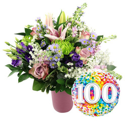 Against a pure white backdrop, a generous celebratory bouquet arranged in a soft, ribbed pink vase captures the essence of a 100-year milestone, the sort often marked in family homes and community spaces around Tufnell Park. The flowers are densely gathered, creating a rounded, luxurious shape brimming with life and colour. Light pink lilies, some fully open with graceful, star-like petals and some still in sleek buds, form the focal point, their presence calm yet impressive. Nestled around them are velvety roses in tones of blush, mauve, and dusty rose, each bloom unfolding with a sense of time and tenderness, like stories passed down over generations. Deep violet lisianthus and small lavender asters with golden centres slip between the larger flowers, adding contrast and the kind of subtle drama that a skilled local florist might bring to a special commission. Crisp lime-green button chrysanthemums punctuate the bouquet, their ruffled forms giving it playful energy. Wisps of pure white baby's breath weave throughout, softening the richer colours and creating a veil-like texture. A mix of dark, glossy foliage anchors the arrangement, suggesting freshness and careful conditioning. Just to the lower right of the bouquet, a shiny round foil balloon gleams, completely covered in bright confetti dots and emblazoned with bold white numerals reading "100," their slight shadow giving a gentle three-dimensional effect. This combination of flowers and balloon feels both elegant and joyful, an ideal tribute for a centenarian, a 100th wedding anniversary celebration, or a historic local business marking a century in the Tufnell Park area, with the design ready to be delivered to nearby streets and cosy venues.