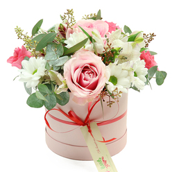 An elegant round hatbox in soft blush pink sits centred against a bright white backdrop, showcasing a carefully curated bouquet that feels both modern and timeless. The box has a velvety, matte texture and is wrapped with a slender coral-red satin ribbon, tied into a neat bow that rests low on the front. A small, rectangular cream-coloured tag hangs from the ribbon, printed with graceful green handwriting and a simple website line, reminiscent of the understated branding you might find in an independent Tufnell Park florist studio near Tufnell Park station. Above the box, the flowers rise in a lush, rounded dome, overflowing just enough to look generous but never untidy. A prominent pale pink rose with a deeper pink heart dominates the centre, its petals open and layered like soft silk folds. Clustered around it are white daisy-like chrysanthemums with bright green centres, their crisp, cheerful faces adding a fresh brightness to the composition. Slender white alstroemeria blooms, each with subtle speckles and fine markings, weave gracefully between the larger flowers, lightening the arrangement and giving it a delicate rhythm. Deeper accents of fuchsia and vivid pink blooms-possibly spray roses or lisianthus-nestle among the pastels, offering lively contrast that lifts the overall palette. Threads of silver-green eucalyptus leaves, with their distinctive rounded shape, mingle with darker green foliage and fine, budded stems, adding both texture and a suggestion of soft, refreshing fragrance. The image evokes the feeling of an intimate, heartfelt gift, the sort of floral hatbox someone in Tufnell Park might have delivered to a Victorian terrace near Carleton Road to mark an anniversary, celebrate a new baby, or simply say "thinking of you" with quiet elegance and care.