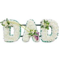 A carefully composed sympathy display that spells DAD in large, three-dimensional letters, each formed from hundreds of small, immaculate white chrysanthemums and tight clusters of button-style blooms to create a plush, uniform surface. The arrangement reads as tactile and calming-the petals appear soft to the touch and the white palette suggests purity and quiet dignity under soft studio light. A vivid emerald-green satin ribbon borders every letter, its sheen sharpening the silhouette and lending a refined contrast to the white massing. Visible behind the letters are dark support bars, indicating the tribute is freestanding and intended to be displayed upright at a funeral, memorial, or wake. Decorative accents are placed with considered restraint: the first D is softened by a delicate cluster of white freesia and fresh green foliage on its upper left, while the last D carries a smaller, complementary freesia and leaf spray on its upper right. Linking the first D and the A are two graceful, arching green stems that sweep diagonally across the composition and terminate in a focused asymmetrical spray at the heart of the display; here two deep purple, almost maroon calla lilies sit amidst white freesias and glossy leaves, their trumpet shapes adding solemn elegance and a touch of dramatic colour. As a Tufnell Park florist familiar with services at nearby Dartmouth Park and along Fortess Road, Flowers Tufnell Park shapes pieces like this to speak plainly of love and loss, suitable for local deliveries and tributes where clarity of message matters.