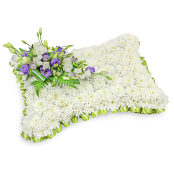 A solemn, lovingly constructed floral pillow composed primarily of tightly clustered white chrysanthemum heads, each blossom contributing to a uniform, cloudlike surface that suggests softness and calm. The shape is a gently rounded rectangle, slightly raised and pillow-like, photographed on a pure white background that accentuates the arrangement's refined lines and the subtle shadow beneath it. Encircling the perimeter is a delicate pleated trim of light green ribbon whose cool, fresh hue frames the white chrysanthemum field and lends a sense of renewal to the tribute. Diagonally across the upper left sits a small, elegant cluster of accent flowers: layers of white lisianthus paired with striking deep purple lisianthus, some in tight bud and some open, their textures and tones evoking dignity and reverent colour. Interwoven are feathery fern fronds and glossy foliage, which lift the cluster with natural movement, while a darker green leaf wrap holds the stems with quiet restraint. The arrangement emits a subtle, floral warmth rather than overpowering scent, designed to comfort rather than distract. As a local florist working in Tufnell Park, Flowers Tufnell Park often crafts this classic pillow for memorials delivered to family homes, chapels along Tufnell Park Road, and small services near Pond Square-an understated, respectful emblem of condolence that balances craftsmanship with heartfelt sentiment.