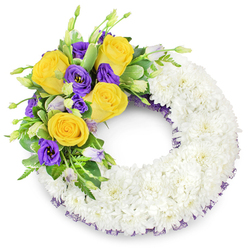 A gracefully constructed circular sympathy wreath, shown against a plain white surface so every detail reads with clarity. The bulk of the design is a continuous ring of fresh white spray chrysanthemums, their tiny multi-petaled heads packed densely to form a smooth, cloud-like texture that suggests calm and unity. Nestled into the upper left portion is a bright, tender spray of flowers: open, sunlit yellow roses whose unfurling petals bring a quiet warmth, contrasted by sumptuous purple lisianthus (Eustoma) with their satin-like, ruffled cups. Scattered among them are a few unopened buds in soft lavender and green, adding a sense of life and gentle progression. The accent is carried by varied foliage-a mix of shiny, rounded leaves, some with faint variegation, and fine fern fronds-that cushions the blooms and gives the composition a natural silhouette. A slender, dark purple mesh ribbon peeks discreetly from beneath the white blooms at both the inner and outer curves, a subtle visual echo of the purple lisianthus. The wreath's open centre and balanced color story of white, yellow and purple make it a fitting tribute for a funeral or memorial, chosen to express sympathy with warmth and restraint. As a local florist might suggest, this piece suits services held around Tufnell Park, Archway and Dartmouth Park where understated beauty and careful craftsmanship are appreciated, and its quiet rose scent and soft textures invite gentle remembrance.