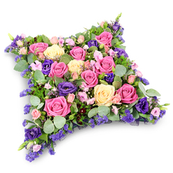 A softly contoured pillow-shaped floral tribute rests against a pure white background, its densely packed flowers forming a reassuring cushion of colour and texture. The shape is a gentle rectangle with rounded corners, suggesting a place where a loved one's head might once have rested, now translated into blooms. Across the entire surface, roses unfurl in varied shades of fuchsia, pastel pink, and creamy pale yellow, their petals layered like satin ribbons, some fully open and lavish, others just beginning to bloom. Threaded between them are deep royal purple lisianthus, the velvety texture of their petals creating pockets of shadow and richness, like quiet pauses in a spoken farewell. Tiny lavender-toned filler flowers, likely statice or limonium, carry wisps of lilac colour through the design, while clusters of blush pink spray roses add a sense of tenderness and intimacy, as if gathered from a much-loved back garden near the tree-lined roads of Tufnell Park. A generous collar of greenery supports the cushion: round, silvery eucalyptus leaves lend a cool, smoky green tone, while darker, more slender foliage adds structure and depth, allowing the brighter flowers to stand out. The overall impression is lush yet serene, with no harsh angles or jarring colours, only a gentle blend of pinks, creams, and purples balanced by fresh greens. The even, neutral light highlights each petal and leaf without distraction, making it easy to imagine this tribute placed at a funeral service, church, or cemetery in and around Tufnell Park, offering a quiet, dignified way to express sympathy, remembrance, and enduring affection when words feel too small.