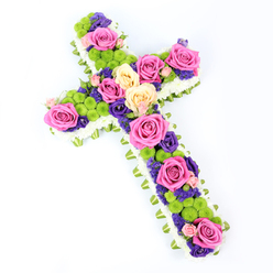 This image presents an elegant floral cross arrangement set against a pure white backdrop, the piece angled diagonally so it appears to gently reach from the upper left of the frame down towards the lower right. The cross is meticulously defined by a pristine edging of small white spray chrysanthemums, each petal forming a plush, cushion-like border that resembles soft clouds around the outer shape. Tucked just beyond this snowy frame, a light green band of folded ribbon or foliage traces the entire outline, adding a fresh, verdant whisper of colour and giving the design a polished, finished look that a local Tufnell Park family might choose for a chapel or graveside service. Inside this border, the cross is densely packed with a harmonious collection of flowers, creating a tapestry of colour and texture. Large hot pink roses and softer creamy yellow or peach roses, fully open and velvety, are positioned with intention along the length and width of the cross, drawing the eye from top to base. Between these classic blooms sit rounded heads of bright lime-green button chrysanthemums, their playful, pom-pom texture breaking up the rose petals and adding a sense of vibrancy. Deeper notes come from smaller blossoms in rich purple tones, such as lisianthus or statice, which nestle into the gaps and lend a quiet depth and formality suitable for a sympathy tribute. The lighting is bright and even, enhancing every subtle shade and curve of the petals without casting harsh shadows. The overall effect is a refined, contemporary funeral flower cross that feels both reverent and gently uplifting, the kind of thoughtful tribute one might order for a memorial in the streets around Tufnell Park Road or nearby Islington crematorium.