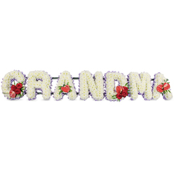 The image shows a lovingly crafted funeral flower tribute spelling out GRANDMA in large, freestanding block letters, presented in a straight row on a clean white backdrop, as if ready to be placed near the coffin at a Tufnell Park memorial. Each letter is formed from tightly massed white cushion chrysanthemums, their small, delicate flower heads pressed closely together so that no gaps of green are visible, resulting in a soft, plush surface reminiscent of snow. Around the outside edge of each character, a narrow trim of pale lavender or soft purple ribbon provides a subtle outline, adding definition and a calm, almost spiritual glow to the white florals. Carefully placed clusters of vibrant red flowers offer moments of colour and emotion: the G holds a group of rich red roses, partnered with a distinctive red-and-white calla lily whose fluted shape arcs gracefully from the cushion of white blooms. The first A is accented with a single heart-shaped red anthurium at its base, its waxy sheen giving a modern yet symbolic touch. At the centre, the M bears another cluster of red roses and a second red anthurium, while the last A mirrors this finishing note with one more anthurium bloom. Beneath the flowers, a dark green support frame discreetly links the letters, giving structural strength without interrupting the design. The tribute feels tender and thoughtful, a personalised farewell arranged by a local Tufnell Park florist, well suited to same-day funeral delivery to chapels and cemeteries around Holloway Road, Archway, and the surrounding North London community.