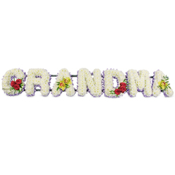 A dignified floral tribute reading GRANDMA stretches horizontally across a clear white background, the simplicity of the setting drawing full attention to the craftsmanship of the arrangement. Each bold capital letter is densely filled with fresh white chrysanthemums, so closely placed that they form a smooth, plush surface, like a blanket of snow. Around the base of every letter runs a slim band of soft lavender or light purple blossoms, creating a gentle halo of colour that frames the word without overwhelming it. Thoughtful flourishes of accent flowers give the tribute a deeply personal feel. On the 'G', three velvety red roses rest on the lower left curve, nestled in dark green foliage that adds depth and contrast. The first 'A' carries two striking chartreuse-green orchids across its central bar, their exotic form hinting at the unique character of the grandmother being honoured. The letters 'R', 'N', and 'D' are kept purely white with the lavender edge, offering a calm, unadorned space that balances the more detailed letters. In the 'M', another trio of rich red roses is tucked into the lower right section, echoing the love expressed on the 'G', while the final 'A' is completed with two more yellow-green orchids to create a harmonious beginning and end. A dark metal frame supports the letters from behind, ready to rest on a stand at a funeral home, crematorium, or family service in and around Tufnell Park, Dartmouth Park, or Holloway Road. The tribute feels serene, heartfelt, and respectful, capturing the tenderness of saying goodbye to a cherished matriarch.