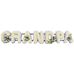 The image shows a horizontal floral tribute spelling the word GRANDPA in large, sculptural capital letters, designed with the care and precision you might expect from an experienced Tufnell Park florist. Each individual letter is completely carpeted in tightly clustered white chrysanthemums, their small, rounded petals overlapping to form a smooth, velvety blanket of blooms. This dense white surface gives the arrangement a soft glow, suggesting innocence, reverence, and a gentle farewell. Along the line of letters, small, intentional bursts of colour appear where rich purple and lavender blossoms have been woven between the white petals, supported by touches of glossy green foliage. These clusters catch the eye at the base of the 'G', in the open spaces of the 'A's, and within the curves of the 'D' and 'P', like little pockets of solace and remembrance. A slim, dark green bar runs beneath the word, providing hidden structure so the letters sit evenly, as if ready to be placed atop a coffin or displayed upon stands during a memorial service in or around Tufnell Park. The calm white and cool violet hues create a sense of stillness that feels at home in a quiet chapel or crematorium chapel nearby, perhaps after a service attended by family from Archway and Kentish Town. You can almost imagine the faint, fresh scent of chrysanthemums in cool air, offering comfort and unity. The tribute's personalised wording, combined with its peaceful palette, makes it a fitting, heartfelt way to honour a beloved grandfather's life and legacy.