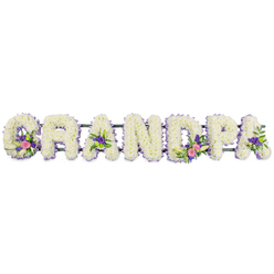 In this photograph, the word GRANDPA is formed from seven substantial floral letters, aligned neatly on a slim dark green bar that holds them in a straight, even row. Each letter is completely covered in compact white chrysanthemums, or similar small multi-petalled blooms, giving the tribute a plush, velvety texture that looks almost like freshly fallen snow. The edges of the letters are trimmed with a delicate border of tiny lilac and purple blossoms, creating a soft halo of colour and gently defining each shape against the clean white background. Scattered through selected letters are expressive accent groupings: the G carries a small posy of white lily-like flowers combined with rich pink and fuchsia tones, the first A and the D show similar clusters of pink, purple, and bright green button chrysanthemums, and another A is touched with coordinated colour and foliage. These accents sit slightly proud of the main surface, adding dimension and visual rhythm without overwhelming the calm white base. The lighting is even and bright, so there are no strong shadows, only the clear, honest detail of each petal and leaf. This is the kind of personalised funeral tribute often ordered for services in and around Tufnell Park, close to Dartmouth Park and Archway, where families want a floral piece that feels both traditional and deeply personal. Although silent, the flowers suggest memories of gentle humour, steady support, and Sunday walks through nearby parks, creating a soothing presence beside the coffin or at the graveside during a final goodbye.