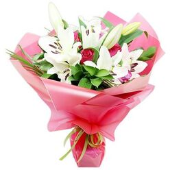 A striking bouquet of white Oriental lilies and deep red roses is shown standing upright against a clean white backdrop, much like a fresh arrangement ready for delivery from a Tufnell Park flower shop. Several large lilies dominate the composition, their elegant, trumpet-like blooms fully open so that each petal curves back softly, revealing the contrasting dark brown, almost aubergine-coloured anthers dusted with pollen. Among these open stars sit a few tall, tapered lily buds, their pale green casings still firm, adding a sense of new life and unfolding beauty. Threaded between the lilies are sumptuous red rosebuds and partially open roses, their velvety petals layered tightly in spirals that give a rich, romantic depth to the bouquet. Surrounding the flowers, generous dark green foliage and fine, grass-like strands fan out, adding a lush, natural framework that enhances the brightness of the white and the intensity of the red. The stems are gathered neatly and wrapped in an ample swathe of shiny, translucent bright pink cellophane, which billows and folds around the blooms like soft fabric, catching highlights in its ripples. At the base, a slim lime green ribbon is tied around the wrapping, its cool, zesty colour offering a modern accent that balances the more traditional flowers. The overall feel is celebratory yet sophisticated, reminiscent of a bouquet you might carry home from a trusted Tufnell Park florist after a stroll along Fortess Road, ideal for birthdays, anniversaries, Mother's Day, or any heartfelt occasion where you want the flowers to speak warmly and clearly.
