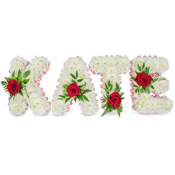 A refined floral name tribute that reads KATE in bold block letters, each letter sculpted from a compact, voluminous swathe of immaculate white chrysanthemums. The tiny petals form a soft, rounded texture reminiscent of linen or fresh snow, and under gentle light the chrysanthemums take on a satin sheen that complements the delicate pale-pink ribbon neatly pleated along every outer edge. The ribbon softens the geometry of the letters and lends a whisper of warmth to the otherwise monochrome field.

Strategically placed, vivid red roses punctuate each letter: the K carries its rose low on the left vertical stroke, the A's red bloom crowns the peak of its triangular form, the T's rose nestles at the base of the left side of the crossbar, and the E is brightened by a rose centered on its middle horizontal bar. Surrounding each rose is an abundant mix of lush green foliage - broad glossy leaves and feathery fern sprigs - that cradle the roses and add natural movement and depth against the white backdrop. The scent is a gentle layering of clean chrysanthemum freshness with the warmer, velvety rose perfume, measured and respectful rather than overpowering.

This piece is crafted with the care of a local Tufnell Park florist who frequently supplies memorial arrangements for nearby churches and community halls. It is designed to convey love, purity, and remembrance in a setting where presence and clarity are paramount.