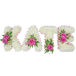 This custom-made sympathy tribute spells out KATE in large, freestanding block letters, each letter built from a dense mass of small, pure white chrysanthemums that create a plush, cloud-like texture. A pale pink satin ribbon delicately outlines the inner shapes of the letters, adding a soft, tactile contrast that guides the eye around the sculpted forms. Vivid hot pink roses are used as concentrated points of colour - two roses on the K, a central rose on the A, one rose anchoring the T, and a pair on the E - their petals unfurling with a gentle scent that lingers above the neutral chrysanthemum base. Lush green foliage, including fern-like fronds, ruscus and variegated leaves, nestles around each rose and peeks from the edges, while tiny light-pink buds add a whisper of additional colour and a garden-fresh detail. Subtle shadows under the letters indicate the arrangement is freestanding and three-dimensional, intended for a memorial table, chapel plinth or celebration-of-life display. The overall impression is tender and well-crafted - a personalised floral tribute meant to convey respect, love and remembrance. Prepared with the care of a local workshop, this style is often supplied for funerals and remembrance services across Tufnell Park and nearby Dartmouth Park; Flowers Tufnell Park can arrange delivery and fitting presentation at local venues and family homes on short notice.