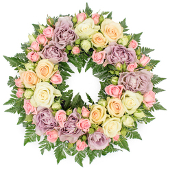 A generously proportioned, circular sympathy wreath is pictured against a spotless white background, allowing every petal and leaf to be clearly seen. The arrangement is richly packed with roses in gentle, muted tones, forming a continuous halo of colour that feels soothing rather than overpowering. Large, open roses in creamy white and soft ivory are dotted all around the ring, their layered petals unfolding in graceful spirals that suggest calm and stillness. Nestled between them, clusters of blush pink spray roses, some just budding and others half-open, give the wreath a delicate, textured appearance, as if capturing many moments in the life of a flower. Hints of pale peach and soft yellow roses bring a quiet warmth, while the more unusual dusty lavender and mauve blooms add depth and a slightly nostalgic character, reminiscent of antique floral prints. Beneath and around the flowers, dark green fern fronds create a lush, feathered backdrop; their fine, pointed leaves emerge from the edges and small gaps, framing the roses and emphasising the circular form. The craftsmanship is meticulous, with each blossom carefully angled so the wreath looks full from every viewpoint, a true florist's piece designed to be seen from a distance at a funeral or memorial. The overall impression is one of gentleness and respect, like a thoughtful tribute placed at a service for a long-loved resident of Tufnell Park, perhaps after a farewell gathering near Tufnell Park station or by one of the quiet side streets leading towards Holloway Road. The wreath speaks in soft colours of remembrance, sympathy, and enduring affection.