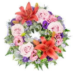 Centred on a crisp white background, a lush circular wreath creates an immediate impression of serene beauty and careful craftsmanship. The form is perfectly round, symbolising continuity, and packed generously with blossoms and foliage so that no gaps disturb the flow of colour. Soft blush and light lavender-pink roses dominate the ring, their fully unfurled petals lending a romantic, almost cloud-like softness. Interwoven among them are lisianthus flowers in shades of pale pink and deep, velvety purple, their ruffled, cup-shaped blooms adding movement and texture. Two large coral to reddish-orange Asiatic lilies command the eye, one near the top arc, the other towards the lower section of the circle, their open faces and visible stamens radiating warmth amid the cooler tones. Wisps of lavender-blue statice or similar filler flowers drift between the larger blooms like a fine mist, tying together pinks, purples, and oranges in a gentle gradient. The greenery beneath is abundant and varied: silvery eucalyptus, slender fern fronds, and glossy deep green leaves give structure and depth, while spiky blue eryngium thistles introduce an unexpected, contemporary note. Unopened lily buds are tucked in among the foliage, suggesting life that carries on and memories still unfolding. A few bright green, folded leaves are artfully arranged near one of the lilies, showcasing skilled floristry. The wreath, which could easily be imagined resting in a quiet Tufnell Park church or accompanying a farewell at a local crematorium, feels like a composed message of peace, sympathy, and unbroken love, created by a florist who understands the gentle, reflective mood of local memorial gatherings.