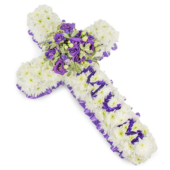 This elegant Christian cross tribute, photographed against a clean white background, is built from a dense layer of white chrysanthemum blooms whose rounded petals create a soft, cushion-like texture across the entire form. A pleated deep purple ribbon trims the perimeter, its ruffled edge giving a stately frame to the design while carrying the colour theme through to the central cluster. Spelling the word "MUM" on the vertical shaft, smaller purple floral heads - likely eustoma or compact spray chrysanthemums - are placed with precision so the letters read clearly from a distance and convey a deeply personal sentiment. The central spray at the arm intersection is a carefully arranged bouquet of dark violet roses and softer lavender lisianthus, their tones moving from dusk to dawn within the same composition; variegated greenery with pale edges and richer centres is tucked throughout to provide contrast and depth, and tiny green berries introduce a tactile, natural element. The light is even and soft, allowing the whites to appear pure and the purples to hold their velvety richness. As a local Tufnell Park florist, Flowers Tufnell Park often prepares pieces like this for services near Tufnell Park Road, Fortess Road, and Dartmouth Park - arrangements meant to express enduring love, comfort, and quiet dignity at a time of farewell.