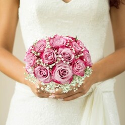 Bathed in soft natural light near Tufnell Park's community gardens, this close-up reveals an exquisite bridal bouquet nestled against the bride's pristine white lace gown. The focus rests on a sumptuous, rounded bouquet of approximately twelve pink roses, each petal unfolding from a pearly, pale centre into rich magenta edges-a subtle gradient reminiscent of a Highgate sunrise. Wisps of pure white baby's breath gently wrap around the roses, forming frothy, cloudlike clusters that lend a lightness to the composition. The bride's hands are carefully poised, nails glazed in the most delicate blush, her warm complexion echoing the gentle energy of a Hertford Road summer. An elegant sleeveless gown is adorned with fine, intricate lace, the V-neckline and flowing sash just visible in the lower right, contributing a further layer of graceful detail. The shallow depth of field draws the gaze exclusively to the tactile beauty of the bouquet-the velvety softness of rose petals set against the feathery texture of gypsophila. One can almost sense the gentle fragrance, floral and uplifting, drifting on the morning air. This image exemplifies the timeless joy and anticipation of a wedding day, and for couples in Tufnell Park, offers a vision of the skilled local craftsmanship and thoughtful design available from neighbourhood florists committed to making each ceremony truly memorable.
