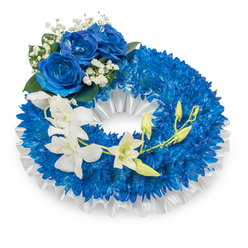 Brought to a calm white setting, this circular sympathy wreath blends a deep blue palette with delicate white accents, an arrangement a neighbourhood florist in Tufnell Park might craft for a local tribute. A voluminous ring of compact, sapphire-blue chrysanthemums creates a richly textured foundation, each tiny, spidery petal contributing to a uniform, velvet-like surface. Lining the inner aperture, a pleated white satin ribbon with a soft, pearly sheen lifts the composition and provides formal contrast to the saturated blue. Anchoring the upper-left quadrant is a trio of large blue roses, fully unfurled and velvety to the eye; their intense azure tones mirror the chrysanthemum base but read as a noble, central cluster. Scattered around those roses are wisps of gypsophila and accents of dark green foliage, which gently punctuate the design with lightness and dimension. A graceful spray of white orchids arcs from the left toward the lower right-Phalaenopsis-like blooms with slender, refined petals that catch a subdued studio light-interrupted by several chartreuse green buds that suggest continuity and hope. There is a faint, cool scent implied by the flowers' textures: the chrysanthemum's mild earthiness, the orchids' faint sweetness, and the powdery hush of baby's breath. As a trained florist servicing Tufnell Park, Dartmouth Park and Archway, Flowers Tufnell Park would present this wreath for a funeral or memorial where understated dignity and enduring remembrance are desired, delivered with attention to condition and timing for funerals or cemetery services.