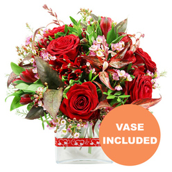 A dramatic, romantic bouquet in rich reds and soft pinks is showcased against a simple white backdrop, allowing every detail of the floristry to shine. At its centre, several sumptuous red roses with velvety petals unfold in layers, some buds still partially closed, others blossoming more fully, capturing that moment just before a rose is at its peak. Around these focal blooms, smaller deep red flowers, much like alstroemeria and similar varieties, cluster in generous groups, adding movement and rhythm to the design. Tiny star-like filler flowers in pale pink and creamy white, similar to waxflower or limonium, are threaded delicately through the arrangement, bringing a light, airy contrast that softens the intense red tones. The bouquet forms a graceful dome of colour and texture, framed by an abundance of verdant foliage. Broad glossy leaves and finer green sprigs intertwine with distinctive burgundy and reddish-brown leaves, some lightly variegated, creating layers of depth that feel as rich and textured as the tree-lined streets around Tufnell Park station and Fortess Road. The flowers are gathered into a clear, contemporary glass vase with a clean rectangular base, allowing the stems and water to be visible and lending the arrangement a modern, uncluttered feel. Around the vase sits a neat red ribbon decorated with repeating white heart outlines, an understated nod to love and affection. In the lower right corner, a prominent circular orange emblem with crisp white uppercase lettering states VASE INCLUDED, indicating that the display arrives complete and ready to place on a table in a Tufnell Park home, perfect for anniversaries, Valentine's surprises, or intimate celebrations.