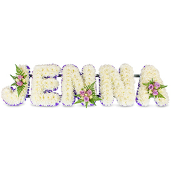 This handcrafted floral composition spells the name JENNA in large, sculpted letters, each formed from a hundred or more small, snow-white chrysanthemums that create a smooth, voluminous surface like soft linen or fresh snowfall. A thin border of purple or lavender-either a delicate ribbon or a fine edge of tiny blooms-hugs the inner contours of every letter, lending a refined silhouette and a whisper of colour that draws the eye along each capital form. Intermittent clusters of spray roses - in subtle blush pinks, pale lavender and a touch of cream - are artfully positioned on key letters: the sweeping curve of the J, the lower centre of the first N, and the upper right of the A. Dark green fern fronds and feathered foliage peek out from these clusters, adding depth, a faint resinous scent, and a natural contrast to the pure whites.

Mounted on slim, dark connecting bars, the five letters read clearly and hold together as a single memorial statement, intended for display at wakes, chapels or celebration-of-life services. Light from a soft morning window would catch the petals and ribbon, creating gentle highlights and a measured sense of hope amid grief. As a local tribute reflecting the calm elegance often requested by families in Tufnell Park and nearby Dartmouth Park, this arrangement balances craftsmanship, restraint and tender colour to honour a name with reverence and quiet beauty.