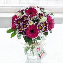 A lush, professionally arranged bouquet is displayed in a squat, clear glass vase, centred against a bright, softly blurred white backdrop that suggests a sunlit window in a calm Tufnell Park home. The arrangement bursts with rich colour and texture: bold gerbera daisies in deep fuchsia and magenta stand proudly at the front, their wide, layered petals surrounding dark central disks that create a striking visual anchor. Around them, bicolour chrysanthemums open like little fireworks, with fringed petals shaded from intense purple at the tips to creamy white near the centre, framing vivid yellow eyes that sparkle with energy. Interwoven among these statement blooms are creamy white spray roses, some unfurling in soft spirals, others still held in compact buds, lending an air of quiet elegance and a hint of sweet scent. Fine sprays of white baby's breath float throughout the bouquet, forming a delicate mist that softens the strong colours and adds a gentle, romantic lightness, reminiscent of early-morning walks along the tree-lined streets between Tufnell Park and Kentish Town. Dense, glossy green foliage forms a supportive base, with leaves curving naturally around the stems, giving the impression of freshly cut garden flowers artfully gathered by an experienced local florist. The vase itself is tied with a simple loop of rustic twine, a small bow resting at the front, from which hangs a rectangular white gift tag decorated with a stylised pink blossom and subtle script, suggesting a handwritten note for a birthday, anniversary, or get-well wish. Soft, even daylight flows across the scene, highlighting the freshness of every petal and the clarity of the glass, evoking the feeling of a same-day flower delivery arriving at a Tufnell Park doorstep, ready to bring colour and joy into someone's living room.
