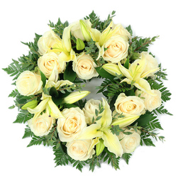 Arranged and photographed flat against a clean white background, this circular funeral wreath pairs creamy white roses with pale yellow lilies, each bloom positioned to create a harmonious, peaceful tribute. The roses, numbering roughly a dozen to fifteen, present compact, ivory-toned spirals of petal that reflect a soft, diffuse light; their surface appears almost luminous beside the matte, lemon-tinged petals of the lilies. Several lilies are fully open with trumpet-like throats and pale stamens visible, while others sit as elegant buds, adding vertical rhythm and the suggestion of future bloom. The base is a lush mosaic of greens-feathery ferns lend a filigree edge, while rounded, slightly greyed-green leaves likely eucalyptus introduce cooler tones and a sturdy textural backbone. From a florist's perspective, the composition balances density and air, the dense clusters of roses offset by the lilies' open geometry and the foliage's directional movement, all arranged to emphasize the wreath's unbroken circle. If you know Tufnell Park, imagine this piece brought gently into a small chapel near Tufnell Park Road or placed at a service in Dartmouth Park: it carries the quiet dignity fitting those neighbourhood spaces. The scent would be subtle-a soft rose perfume brightened by green, citrus-suggestive notes-and the palette reads as both elegant and consoling. Prepared with careful craftsmanship and a restrained hand, this wreath offers a respectful, timeless expression of sympathy suitable for funerals, memorials, or as a home remembrance.