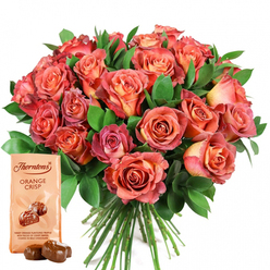 Arranged with the seasoned touch of a florist from the heart of Tufnell Park, this bouquet of roses radiates warmth and sentiment as it graces a spotless white canvas. The collection is alive with subtle, shifting hues-petals unfurl from burnished coral centers to gentle peaches and blush pinks at their edges, each bloom a glowing lantern cradled by the glossy, deep greenery of eucalyptus or salal. The interplay of colour and form suggests the gentle haze of an evening sun over Dartmouth Park, with every flower promising a tactile pleasure, from velvety petal to smooth leaf. The bouquet, reminiscent of late summer's golden hours, pairs effortlessly with an inviting packet of Thorntons Orange Crisp chocolates at its base. The soft cream wrapper bears the Thorntons name, and two glossy chocolate truffles lie temptingly in the foreground, suggesting sweetness inside and out. This artful offering, designed for birthdays, anniversaries, or spontaneous celebrations, brings together the romance of roses and the indulgence of chocolate. Perfect for residents along Fortess Road or those marking milestones in local cafés, the set is not just a visual delight but a multisensory gift that echoes the community spirit of Tufnell Park.