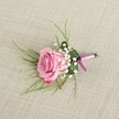 A single pink rose boutonniere lies on a softly textured, light beige linen cloth, captured as if viewed from just above a jacket lapel, the kind of detail you might notice on a groom stepping out of a town house near Tufnell Park underground. The rose itself is in a semi-open bloom, its velvety petals spiralling from a gentle blush at the heart to a slightly deeper pink at the edges, suggesting both freshness and quiet romance. Around its base, tiny clusters of white baby's breath form a light, cloud-like halo, each minuscule blossom adding a dusting of softness that contrasts with the smooth curves of the rose. From behind the central flower, fine, grass-like strands of bright green foliage splay outward in delicate arcs, while a single broader, darker green leaf is neatly folded to cradle the stems, giving the small arrangement a sense of structure and polish that would suit any elegant wedding in Tufnell Park or a prom at a nearby North London school. The stems are carefully wrapped in a satin ribbon that blends lavender-pink with hints of deeper magenta, tied in a simple, secure knot that catches the light just enough to feel refined rather than showy. The woven linen background, with its subtle, natural texture, provides a calm, neutral stage that lets the pinks, whites, and greens stand out clearly. Gentle, even lighting falls across the scene, creating soft shadows that reveal the three-dimensional shape of the boutonniere and the craftsmanship behind it, making this image an ideal visual for anyone imagining classic, romantic floral details for a formal celebration near Tufnell Park's leafy streets.