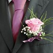 A poetic arrangement unfolds in this Tufnell Park boutonniere, every element chosen with a sense of romance and place. At the composition's heart rests a partially opening pink rose-its petal blush alive with softness and hope, a nod to the optimism found in local vows and promises. The delicate fragrance-implied but nearly tangible-suggests fresh mornings shared in the leafy walks of Tufnell Park itself. A halo of baby's breath encircles the rose's gentle core, their snowy white blossoms likened to the morning mist that often settles along residential lanes and the expanses of Tufnell Park Gardens. Slender arches of deep green foliage and a discreet fan of broad leaves give shape and vitality, lending depth and grace reminiscent of a well-loved local park after rain. Anchoring the design, a trailing satin ribbon in lavender tones echoes the lilac-pink tie neatly knotted atop the wearer's light, subtly patterned grey shirt. The boutonniere is secured on the lapel of a dark grey suit jacket, its textured weave and lapel detail inviting association with the refined yet accessible fashion seen at Tufnell Park's community events. Ideal as inspiration for special occasions-weddings, proms, or life's cherished milestones-this floral accent crafted by Flowers Tufnell Park captures more than beauty; it adds meaning, artistry, and a local sense of belonging to every formal attire.