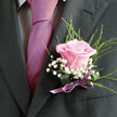 The image offers an intimate, chest-up view of a man dressed for a special occasion in Tufnell Park, drawing the eye straight to the refined details of his suit and boutonniere. His jacket is a dark charcoal grey, almost black, with a cleanly pressed lapel on the left where the flowers are pinned. Underneath, a light dress shirt with a delicate gingham or checked pattern peeks out, its cool tones setting off a vivid magenta tie that runs smoothly down the centre of his torso. The tie's satiny sheen catches the light, suggesting early evening or soft indoor lighting, like you might find in a townhouse wedding off Junction Road. Nestled securely on the lapel, the boutonniere features a single pale pink rose, its petals soft and full, opening in a perfect spiral from the deeper pink heart to softly blushed, almost translucent edges. Clusters of white baby's breath surround the rose, tiny star-like flowers forming a misty border that makes the central bloom stand out with romantic clarity. Fine, bright green strands of foliage rise behind and around the blossoms, giving the composition a sense of airy height and a freshly picked feel, as though it had just been arranged by a careful local florist. The stems are wrapped in a smooth deep green band and finished with a slim purple ribbon tied into a neat bow right above the breast pocket. The ribbon's muted lavender note harmonises with the pink rose and tie, creating a balanced, modern colour story that quietly conveys affection and celebration, ideal for weddings, proms, or formal gatherings across this corner of North London.