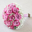 A romantic bouquet of roses and baby's breath is displayed on a soft, cream-toned textile background, photographed as if resting on a table in a calm Tufnell Park living room. The arrangement is positioned on a diagonal, giving a sense of movement, with the flower heads fanning towards the upper left and the stems tapering towards the lower right. At the centre, approximately a dozen plump roses create a lush, rounded cluster. Their colouring is quietly dramatic: the innermost petals are a gentle, dusty lavender-pink, gradually intensifying into bright fuchsia and deep magenta at the outer curl of each petal. This gradient gives every rose a painterly depth, as though brushed with layers of spring evening light. Some blooms are still modestly closed, their edges touching in tight spirals, while others open more generously, revealing the intricate whorls typical of freshly cut, high-quality roses. Surrounding them is a generous froth of white gypsophila, its tiny blossoms resembling airy confetti floating between the larger flowers. The baby's breath creates a soft, cloudlike border, offering visual breathing space and enhancing the vibrancy of the pink tones, much like the quiet contrast between leafy side streets and the bustle of nearby Kentish Town. A few rich green leaves tuck in around the base of the blooms, lending natural contrast and a garden-fresh feel. The stems are bound in a smooth, lilac satin ribbon, carefully wrapped to create a sleek, comfortable handle. A delicate spiral of small white pearls is pinned into the ribbon, catching the light and echoing traditional bridal jewellery. The bouquet feels ideal for a wedding, engagement celebration, or special occasion dinner hosted in one of Tufnell Park's elegant Victorian homes, capturing the emotion of devotion and cherished promises.