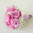 This vibrant, hand-tied bouquet captures the essence of Tufnell Park floristry at its finest, marrying boldness and delicacy in a compact, celebratory arrangement. Five full, beautifully curled roses shine at the centre, their petals transitioning from dusky lavender at the base to the striking deep-pink edges, each bloom appearing fresh and full of life. Dainty white baby's breath clusters are expertly placed between the roses, lending an airy softness that seems to float above the textured, pale beige linen surface below- reminiscent of the welcoming homes around Lady Somerset Road. The neatly gathered stems are bound in a glossy magenta-plum ribbon, textured with a subtle shimmer and enhanced by parallel spirals of lustrous pearls, forming an elegant, tactile handle. At the base, the natural green ends of the stems are visible, a nod to the bouquet's local, freshly-sourced appeal. Gentle daylight plays across the ensemble, illuminating every intricate curl and reflecting off the pearled ribbon. Perfect as a bridesmaid's accessory or as a charming centerpiece for a wedding breakfast in Tufnell Park, the bouquet radiates warmth and joy, capturing both the romantic symbolism of roses and the airy lightness of gypsophila. This arrangement reflects the artistry and care of Tufnell Park's local florists, offering inspiration and elegance for any intimate celebration in North London.