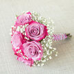 On a pale beige, subtly woven fabric background, a compact bouquet lies diagonally as though resting before being handed to its owner, its colours vivid yet harmonious. The focus is a cluster of luxurious, bi-colour roses, each one unfurling from a cool, lavender-tinged heart into outer petals edged with a lush, dark fuchsia and magenta pink. This gradual shift in shade gives every bloom a sense of movement, like a gentle swirl of colour held in mid-air. Between these roses, sprays of white baby's breath-gypsophila in tiny, rounded clusters-float like wisps of cloud, softening the intense pink tones and lending the arrangement an airy, timeless charm. The florist has bound the stems with a band of satin ribbon in deep purple, its sheen catching the light in narrow highlights that hint at silky texture beneath the fingers. Over this ribbon, a string of small, white faux pearls is wrapped in a measured spiral, each bead sitting neatly, as if patiently placed one by one to create a subtle, vintage-inspired luxury. The bouquet feels perfectly sized for a bridesmaid walking through Tufnell Park's leafy streets to a ceremony, or for a prom guest stepping out near Tufnell Park station. Although there is no visible greenery beyond the stems, the interplay of magenta, lavender, and white against the calm neutral background creates a balanced, contemporary look. The image conveys the impression of fresh, carefully conditioned flowers, likely arranged the same day by a local Tufnell Park florist for delivery to a home near Brecknock Road or a reception space overlooking Parliament Hill Fields.