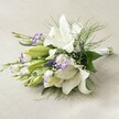 Lying gracefully across a textured, pale linen surface reminiscent of a Tufnell Park townhouse interior, this hand-tied bouquet is a testament to both classic elegance and the neighbourhood's understated charm. Large, pristine white Oriental lilies, their trumpet-shaped blooms fully open or just beginning to unfurl, form the heart of the arrangement. Shimmering yellow anthers and delicate speckles in the blooms' throats catch the gentle natural light, evoking purity and celebration. Interlaced between the lilies are smaller, enchanting Lisianthus-each bloom sporting white, ruffled petals with striking purple edges, adding depth and comfort much like evenings spent strolling along Fortess Road. Slender green stems support clusters of tiny purple florets, perhaps Veronica, which contribute vertical lines and richness. Whisper-light sprays of baby's breath dance throughout, lending softness and a touch of romance, while verdant, feathery foliage and deep green leaves hug the bouquet's base and spill gently outward. All stems are neatly gathered with a hint of white ribbon, reflecting the careful craftsmanship local to Tufnell Park's community florists. The composition radiates purity, hope, and a quiet sophistication-perfect for bridal parties or treasured moments in this north London area. Each element, highlighted by even lighting, celebrates both nature's artistry and human care, making this bouquet a heartfelt choice for special occasions.