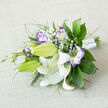 Arranged with a thoughtful, slightly asymmetrical hand and pictured on a neutral, woven linen surface, this bouquet reads as a study in elegant contrasts. Large white Oriental lilies provide a luminous focal point: some petals unfurl fully to reveal warm yellow-orange stamens and delicate dark flecking, while other buds remain a fresh, vibrant green, promising to open. Interspersed are lisianthus (eustoma) blooms: their soft white centres melt outward into vivid lilac and deep purple fringes, the ruffled edges recalling hand-painted porcelain. Wisps of baby's breath breathe between the larger flowers, lending an airy, whimsical cadence that catches the light like tiny, floating clouds. The greenery - broad, glossy leaves likely salal or ruscus, coupled with finer, feathery fronds and spiky ornamental grasses - provides depth, texture and a cool palette of greens that anchors the composition. The stems are bound in white satin ribbon, discreetly embellished with pearl-like studs for a refined finish that still feels intimate. Photographed in soft daylight, the bouquet's textures - waxy lily petals, crimped lisianthus edges, and matte linen - are all clearly legible to the eye and suggest a gentle fragrance. As a Tufnell Park florist, we imagine this bouquet arriving for a small ceremony near Fortess Road or a wedding breakfast in Dartmouth Park; it carries a calm, celebratory mood suited to bridal parties, anniversaries, or a thoughtful gift to mark a new chapter, available for careful local delivery across Tufnell Park and neighbouring Kentish Town.