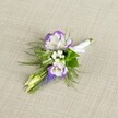 An elegantly crafted wedding boutonniere lies carefully placed on a softly textured light beige linen background, the neutral fabric recalling the calm interiors of townhouses along Tufnell Park Road. At the heart of the piece sit two open lisianthus blooms, their petals a pure, velvety white delicately ruffled and traced with a deep, vibrant purple edge, like ink touched lightly to paper. Nestled close to these central flowers are airy clusters of tiny white filler blooms, similar to baby's breath, which give the buttonhole a cloudlike softness and a sense of lightness against the warm, woven cloth. Threaded through the composition is a slender vertical spike of tightly packed, bell-shaped muscari in a rich, grape-purple tone, adding a touch of woodland charm that feels at home near Tufnell Park's leafy side streets and nearby Hampstead Heath. Fine, feathery green foliage, reminiscent of asparagus fern, fans gently around the flowers, its soft texture creating movement and framing the blooms with a fresh, garden-picked feel. Two pale green, unopened buds peek from the design, suggesting new beginnings and the quiet anticipation of the day ahead. All the stems are drawn neatly together and wrapped in a smooth white satin ribbon, secured in a refined finish that signals this piece is ready to be pinned to a lapel. The overall mood is calm, romantic, and understatedly sophisticated, suitable for a groom or groomsman preparing for a ceremony in one of Tufnell Park's intimate venues, or for anyone wanting a floral accent that whispers elegance rather than shouts for attention.