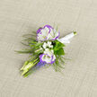 A small composition with quiet presence, this boutonniere pairs two ruffled lisianthus - white at their centers and edged in a gentle violet - with a sprinkling of baby's breath that softens the overall silhouette into a cloud-like halo. Pale, unopened buds extend as slender murmurs around the main blooms, their shapes suggesting fresh energy and restraint at once. Below, compact spikes of grape hyacinth in a vivid blue-violet provide a jewel-toned contrast that draws the eye downward, balancing the airy whites above. Delicate ferns arc around the arrangement, introducing a woodland texture that feels both fresh and timeless. The stems are bound in a small white ribbon, the knot tidy and understated, and the boutonniere is pictured on neutral linen which highlights the cool palette and the flowers' satiny surfaces. In the softer afternoon light typical of Tufnell Park's garden squares, the colours take on a tranquil, almost painterly quality. As a local florist I think of this piece as perfectly suited to the lapel of a tailored jacket or the buttonhole of a long dress - a subtle punctuation of style at weddings, milestone celebrations, or formal portraits. At Flowers Tufnell Park we often assemble boutonnieres like this when couples ask for seasonal, textured accents that feel personal to the neighbourhood and quietly memorable.