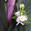 A close-up portrait of elegant wedding attire shows the upper torso of a man dressed for a special celebration in Tufnell Park. He wears a sharply tailored charcoal grey suit jacket with clean, structured lines and a soft sheen that suggests fine fabric, layered over a neat light grey checkered shirt. At the centre of the frame, a lustrous magenta silk tie catches the light, its rich colour echoing the joyful mood of a wedding morning near Tufnell Park station. Pinned to the left lapel is a carefully crafted boutonniere: a small, exquisite cluster of white lisianthus blooms rimmed with deep purple, their ruffled petals opening gently around bright yellow centres. Around them, slender white buds hold the promise of flowers yet to open, while a single vertical spire of tiny, dark violet veronica florets adds height and quiet drama. Wisps of feathery green foliage and smooth, broader emerald leaves weave through the design like a tiny garden, softened by airy flecks of baby's breath. A fine loop of decorative green grass adds a modern flourish, as if the florist has drawn a graceful line through the air. At the base, a pristine white satin ribbon is tied into a neat bow, grounding the flowers with a touch of tradition. The overall effect is one of poised, understated romance-a groom or groomsman ready to step into a ceremony, perhaps in a townhouse venue off Brecknock Road, wearing a piece of living artistry close to his heart.