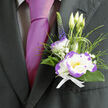 This boutonnière has the composed authority of a piece crafted by a neighbourhood florist who knows the rhythm of Tufnell Park mornings. At its centre sits a white lisianthus, its petals tipped in vivid purple that provides a fine-lined definition as light skims the bloom; alongside it, a compact white dianthus with playful purple ruffles introduces soft, tactile interest. Small clouds of gypsophila breathe lightness into the arrangement, while a singular, upright indigo veronica spike lends height and an unexpected, elegant note. Feathery ornamental grass and dark green foliage form a natural cradle, their cool textures and slender movement suggesting fresh-cut stems from a local market stall on Tufnell Park Road. Everything is secured with a crisp white ribbon, neat and purposeful, and placed against a dark suit lapel next to a magenta tie for a striking but tasteful contrast. The overall effect is sophisticated yet approachable-the sort of piece Flowers Tufnell Park would prepare for a groom, an honoured guest, or anyone seeking a refined floral accent for a wedding or anniversary. Soft garden-scented notes accompany the wearer, comforting and subtle through photographs, vows, and evening toasts. This boutonnière balances classic formality with a wink of personality, crafted to endure a long, joyful day while echoing the calm confidence of the local neighbourhood.