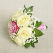 Laid at a diagonal on a pale beige woven background that resembles fine linen, this compact bouquet is a study in considered contrast and graceful craftsmanship. Five creamy ivory roses form the central cluster, each bloom opening into plush, layered petals that diffuse the soft studio light and suggest a gentle, powdery rose scent. Interlaced among them are delicate clouds of Gypsophila-baby's breath-whose tiny white blossoms float around the roses like soft mist, adding air and lightness. Off to the front-left, a single fuchsia-pink rose asserts itself with vivid cheer, its richer tone creating a striking focal point amid the neutrals. Lush, pointed green foliage-probably ruscus or salal-peeks through, giving depth and a fresh, leafy coolness to the arrangement. The stems are carefully bundled and wrapped in a bright pink satin ribbon that forms a smooth, tactile handle; several strands of small, iridescent white pearls are laid across the ribbon in neat parallel rows, catching the light and lending a timeless, bridal elegance. Captured from a close, slightly elevated angle with even, flattering light, the image highlights texture-the satin's sheen, the roses' velvet, and the background fabric's weave-inviting touch. I craft pieces like this often for weddings and proms around Tufnell Park, and at Flowers Tufnell Park we take care to balance colour, scent, and scale so a petite posy can feel utterly memorable and right at home near Tufnell Park Road or the tube station.