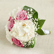 Displayed on an off-white, slightly textured fabric that feels like natural linen, this bouquet captures the refined yet relaxed charm of a Tufnell Park wedding morning. The eye is immediately drawn to a large, fully open peony in creamy white, placed centrally and slightly forward, its petal edges soft and feathery, as though gently lifted by a breeze. Surrounding it, several fuchsia-pink roses nestle close, their classic spiral centres still tight and full, embodying romance and anticipation. Tiny white stars of gypsophila thread their way throughout the composition, forming a cloud-like veil that lightens the overall look and adds a sense of tenderness. Around the base, wide, dark green leaves arc elegantly, their rich colour forming a graceful frame and echoing the greenery of nearby Tufnell Park gardens and tree-lined streets. The flower stems are gathered into a neat bundle and wrapped with smooth white satin ribbon, wound carefully in even bands to create a clean, sophisticated handle, suitable for a bride walking into a ceremony at a local church or intimate venue. The lighting is soft and natural, like filtered daylight through a townhouse window, highlighting the variations of cream, pink, and green without creating harsh contrast. Texture plays a central role: the plush peony petals, the firm rose blooms, the airy gypsophila, and the sleek foliage together create a design that feels both luxurious and approachable. This bouquet would suit spring or summer celebrations anywhere around Fortess Road or Dartmouth Park, evoking serenity, affection, and timeless elegance.