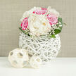 In this softly lit scene, a lush bouquet of creamy white peonies and vivid fuchsia-pink roses is arranged in a perfect rounded dome, evoking the grace of an intimate Tufnell Park wedding or a romantic dinner just off Fortess Road. The peonies sit at the heart of the design, their many velvety petals layered like satin, forming generous blooms that appear almost weightless in the bright, even light. Around them, pink roses glow with deeper magenta tones towards their centres, some petals gently curling at the edges as though just beginning to open to the day. Wisps of baby's breath weave between the larger flowers, their tiny white blossoms giving a gauzy, cloud-like softness, as if a light veil had been draped over the design. Rich green leaves circle the base, their smooth surfaces catching subtle highlights and shaping the bouquet into a classic, compact bridal-style cluster. The flowers are gathered in a striking white vase fashioned from interlaced, branch-like strands that form an open, basket-like structure, its airy design offering glimpses of stems and creating intriguing shadows. In the foreground, resting on a clean, white surface, two pale woven balls echo the vase's texture, like small pieces of decor you might spot in a thoughtfully styled living room near Tufnell Park station. The backdrop is a neutral, linen-textured beige, calm and understated, allowing every petal and leaf to take centre stage. The overall feeling is one of poised elegance and hopeful beginnings, ideal for marking milestones, anniversaries, or sending a heartfelt message with same-day delivery across Tufnell Park and nearby Kentish Town.
