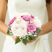 A romantic bridal bouquet rests gently between a bride's hands, its compact, rounded shape built around opulent ivory peonies and pastel roses, like something carried from a ceremony on a side street near Campdale Road in Tufnell Park. The peonies are the stars here: large, cloud-like blooms in the palest cream, their many silken petals layered so densely they appear almost weightless, suggesting a softness you can almost feel. Nestled among them are roses in harmonious shades of dusty pink and lavender, each rose opening in a slow spiral, creating a sense of poised calm rather than showiness. Fine sprays of gypsophila weave in and out of the main flowers, like a dusting of delicate snow or lingering spring blossom, lending a subtle, airy texture that keeps the bouquet from feeling too heavy. Framing everything are rich green leaves with a sturdy yet elegant presence, their deeper tone grounding the pastels and giving the design a classic bridal profile. The stems appear to be carefully gathered and likely wrapped in a simple ribbon, leaving the emphasis entirely on the flowers themselves. Gentle daylight, maybe from a large Victorian window in a Tufnell Park townhouse, picks up the velvety texture of the petals and the slight sheen of the foliage, making the whites more luminous and the pinks more tender. This bouquet feels tranquil and thoughtful, the kind of arrangement a local florist might create for a bride who loves understated beauty, fresh seasonal blooms, and a wedding day filled with quiet joy rather than grand spectacle.