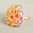 Captured from just above and resting horizontally on a woven, light beige backdrop, this elegant bouquet is an intimate study in warm tones and soft textures. The central form is a compact, rounded bouquet of fully opened roses in peach and soft orange shades; their petals curl outward in layered spirals, creating a lush, tactile surface that seems almost velvety to the fingertip. Threaded between those roses are crisp white lisianthus whose ruffled edges are tipped with vivid violet, a detail that brings depth and a touch of playful drama to the arrangement. Tiny sprays of gypsophila-baby's breath-float between the larger blooms, their tiny white buds like pinpricks of light that add an airy counterpoint to the dense rose heads. The bouquet's handle is wrapped in a peachy coral satin ribbon, and a string of creamy, off-white pearls spirals elegantly around the stems, introducing a whisper of vintage glamour. Soft, diffused daylight softens the scene and highlights subtle colour gradations while the textured fabric keeps the focus on craftsmanship. As a local florist familiar with weddings around Tufnell Park Road and intimate gatherings near the tube station, I picture this bouquet in the arms of someone walking from the station toward a neighbourhood celebration-its scent of fresh roses trailing behind. The overall mood is romantic and refined, a design that balances tradition with a modern twist and feels perfectly at home in this corner of north London.